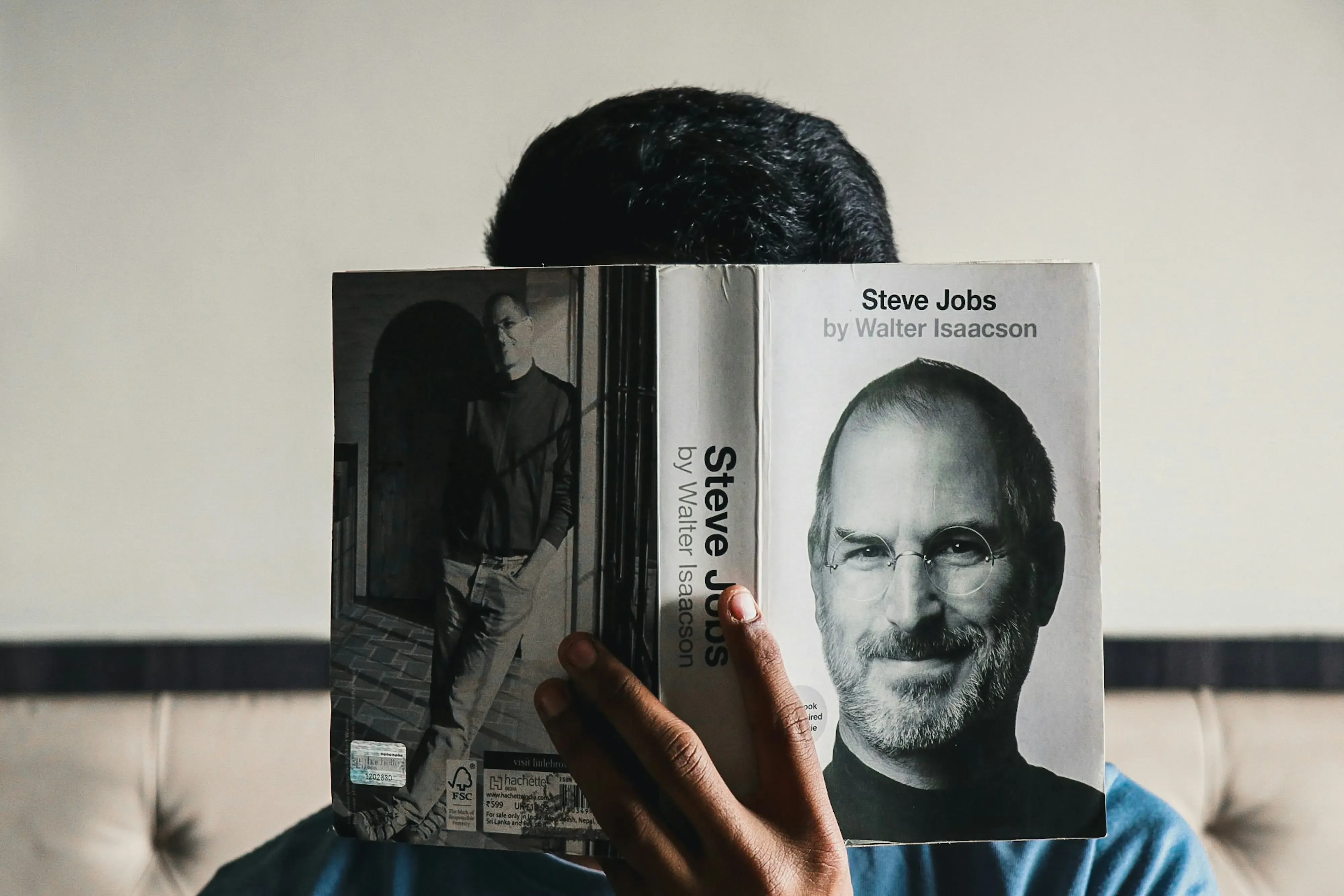 A person holds the biography 'Steve Jobs' by Walter Isaacson in front of their face, obscuring their features, with Steve Jobs' portrait on the book's cover looking directly at the viewer