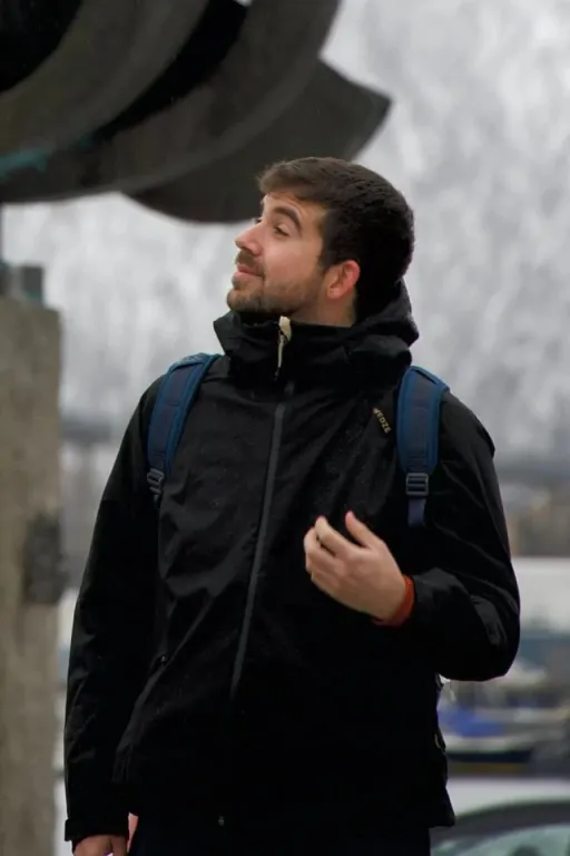 Me looking left with a smile. Image was taken in Tromsø, Norway. I'm wearing a black jacket and a blue backpack.